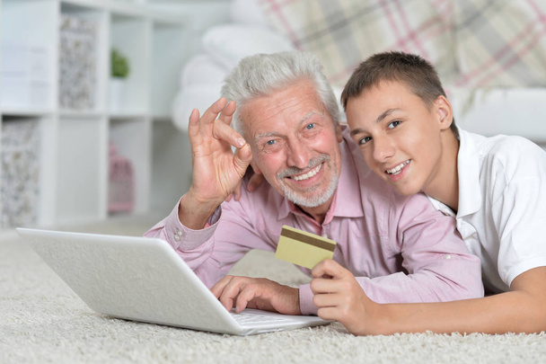 abuelo y nieto acostado en el suelo y el uso de ordenador portátil, nieto con tarjeta de crédito, concepto de compras en línea
 - Foto, imagen