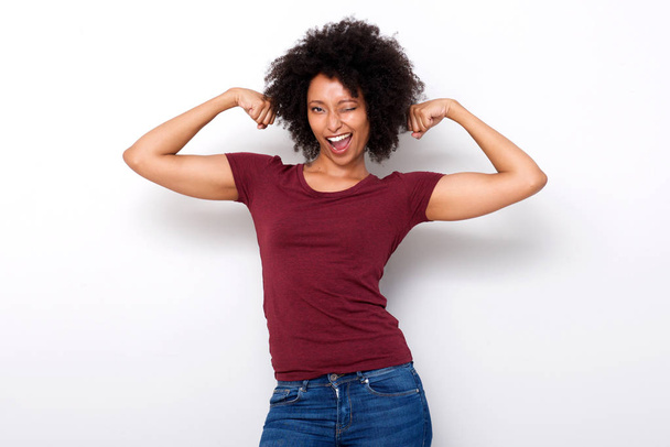 Portrait de forte jeune femme africaine fléchissant les deux muscles des bras et clignant des yeux sur fond blanc
 - Photo, image