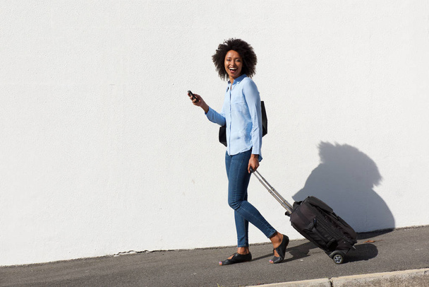 Full body portrait of laughing young female traveler walking with suitcase and mobile phone - Photo, image