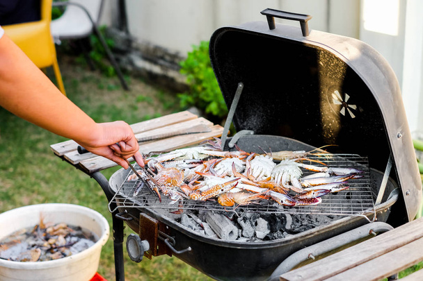 Hand holding silver tongs for picking crab & shrimp (seafood) and burn with black charcoal on metal grid - party food & picnic concept in the summer. - Photo, Image