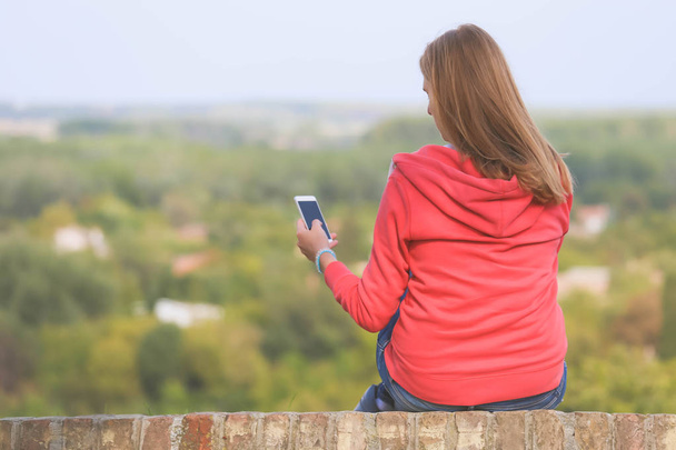 Το κορίτσι που κάθεται σε ένα τοίχο που αντιμετωπίζει την πλάτη της και χρησιμοποιώντας το smartphone - Φωτογραφία, εικόνα