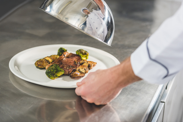bijgesneden afbeelding van chef-kok plaat met maaltijd en deksel houden op restaurant keuken - Foto, afbeelding