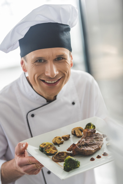 гарний шеф-кухар тримає тарілку з вареним стейком на кухні ресторану
 - Фото, зображення