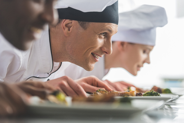 hymyilevät monikansalliset kokit haistelevat keitettyä ruokaa ravintolan keittiössä
  - Valokuva, kuva