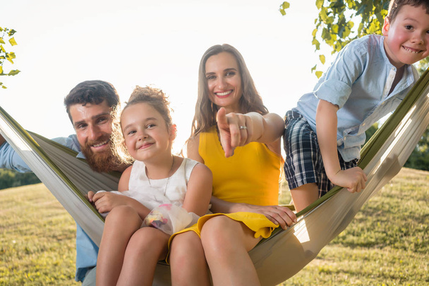 Family portrait with a beautiful mother of two playful children swinging in a hammock while looking at camera next to her husband outdoors in summer  - Foto, afbeelding