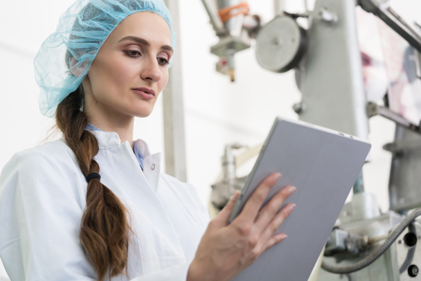 Female expert wearing white lab coat and protective headwear while analyzing information on a tablet PC during work in a contemporary factory - Φωτογραφία, εικόνα