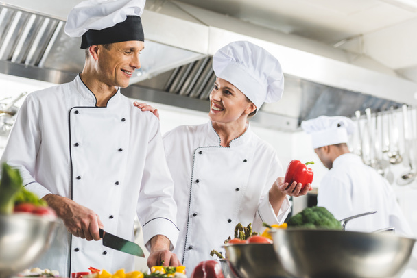 σεφ δείχνει πιπεριά και palming συνάδελφο ενώ αυτός κοπής λαχανικών σε κουζίνα εστιατορίου - Φωτογραφία, εικόνα
