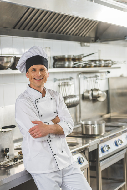 smiling chef standing with crossed arms and looking at camera at restaurant kitchen - Fotografie, Obrázek