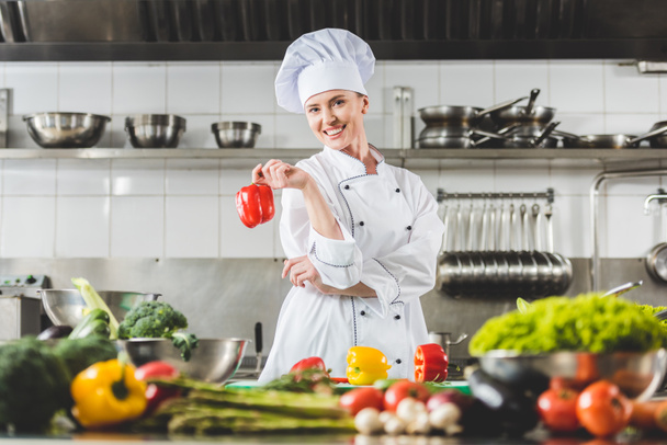 smiling chef holding red bell pepper and looking at camera at restaurant kitchen - Foto, imagen