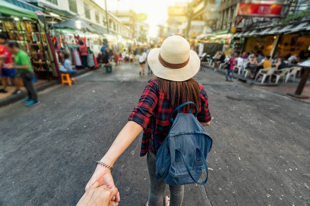 Back side of Young Asian traveling women leading by holding her boyfriend hand in Khaosan Road walking street in evening at Bangkok, Thailand, traveleling together and tourist with follow me concept - Photo, Image
