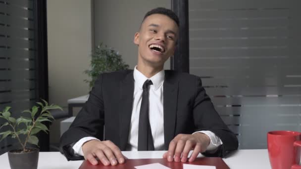 A young successful African businessman in a suit is sitting in the office and laughing while looking at the camera. Gestures with his hands. 60 fps - Séquence, vidéo