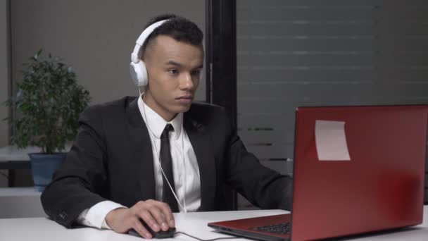 Young successful African businessman in a suit is working on a computer in headphones, playing a computer game at work. 60 fps - Video, Çekim