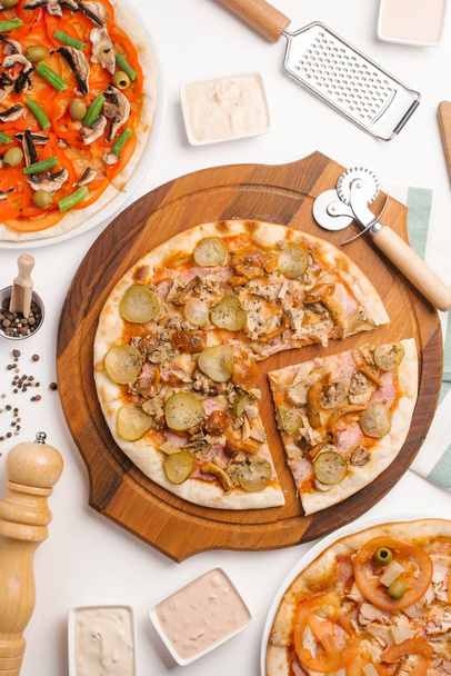 Top view of different kinds of pizza served on wooden pizza plate and white plates, pizza cutter, grater, sauces, pepper grinder, scoop and napkin on white table - Photo, Image