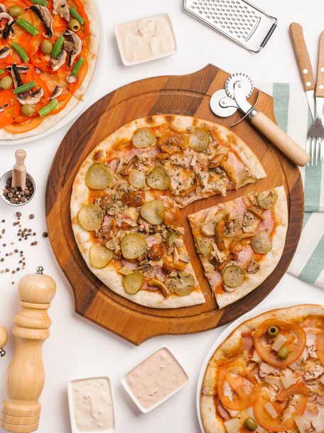 Vue rapprochée de trois types de pizza servis sur table blanche avec sauces, serviettes et ustensiles de cuisine
 - Photo, image