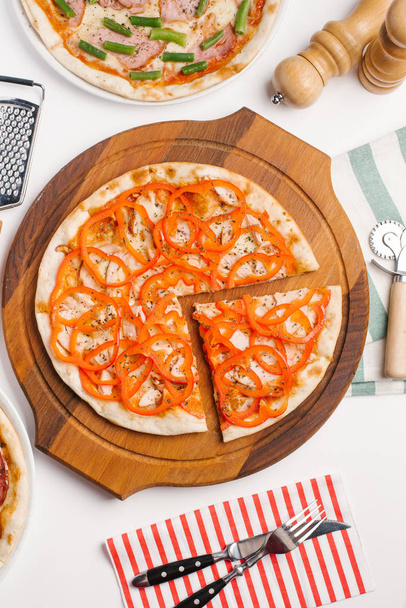 Vue de dessus de différents types de pizza servis sur une assiette de pizza en bois et des assiettes blanches, coupe-pizza, râpe, sauces, moulins à poivre et sel, râpe et serviette sur table blanche
 - Photo, image