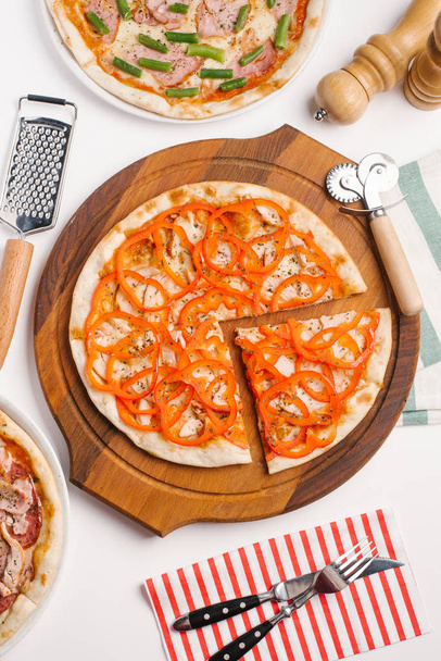 Vista superior de diferentes tipos de pizza servida en plato de pizza de madera y platos blancos, cortador de pizza, rallador, salsas, molinillos de pimienta y sal, rallador y servilleta en mesa blanca
 - Foto, Imagen
