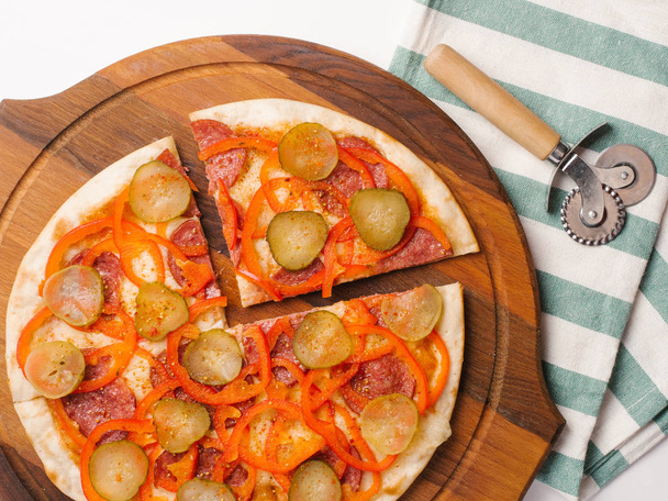 Vista cercana de la pizza en rodajas con salsa de tomate, queso mozzarella, pepperoni, pimientos dulces y encurtidos servidos en un plato de pizza de madera con cortador de pizza y servilleta
 - Foto, Imagen
