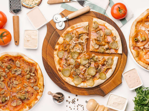 Vue de dessus de trois types de pizza servis sur une table blanche avec sauces, légumes, serviettes et ustensiles de cuisine
 - Photo, image