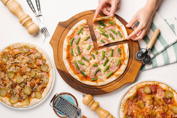 Mãos masculinas tomando pedaço de pizza sentado na mesa branca com diferentes tipos de pizza, faca, garfo, cortador de pizza e guardanapo
 - Foto, Imagem