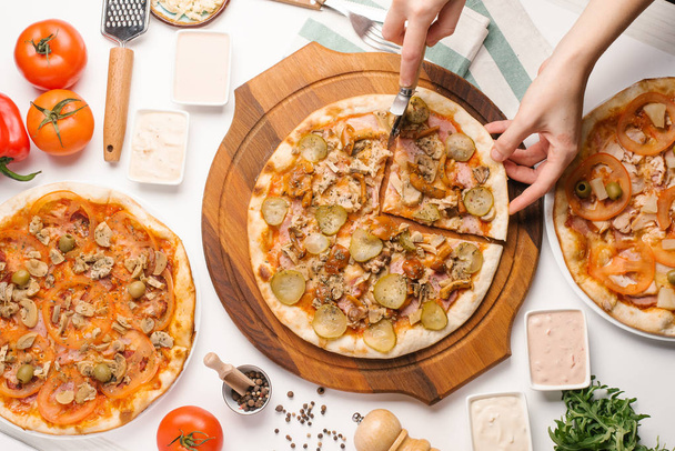 enge Ansicht weiblicher Hände, die Pizza mit Pizzaschneider mit verschiedenen Arten von Pizza, Soßen, Gemüse und Geschirr auf weißem Tisch schneiden - Foto, Bild
