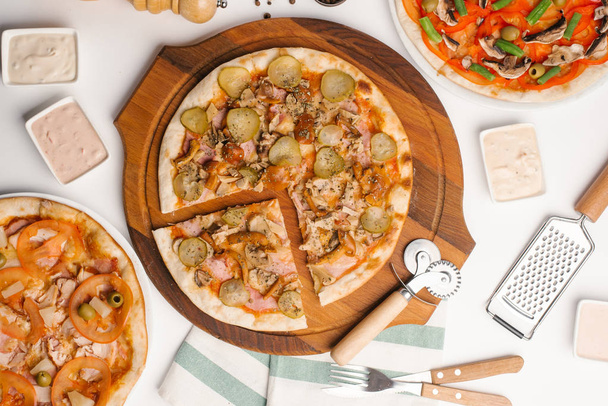 Vue de dessus de trois types de pizza servis sur une table blanche avec sauces, serviettes et ustensiles de cuisine
 - Photo, image