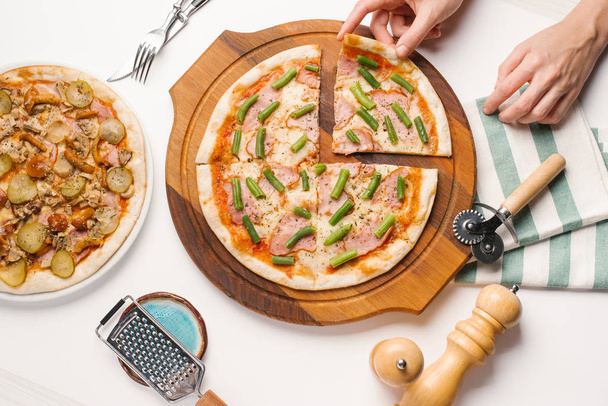 Widok z góry męskich rąk biorąc kawałek pizzy, siedząc przy stole biały z różnych rodzajów pizzy, nóż, widelec, nóż do pizzy i serwetka - Zdjęcie, obraz