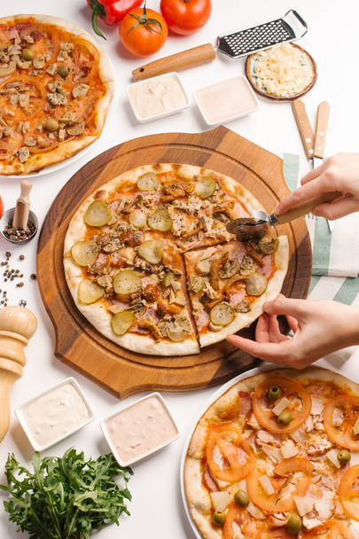 Vue rapprochée des mains féminines coupant la pizza avec coupe-pizza avec différents types de pizza, sauces, légumes et ustensiles de cuisine sur table blanche
 - Photo, image