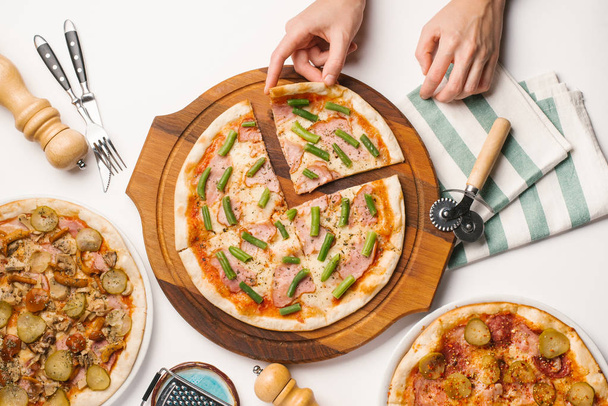 Closeup mužských rukou, přičemž kus pizzy sedí u stolu bílý s různými druhy pizzy, nůž, vidličku, kráječ na pizzu a ubrousek - Fotografie, Obrázek