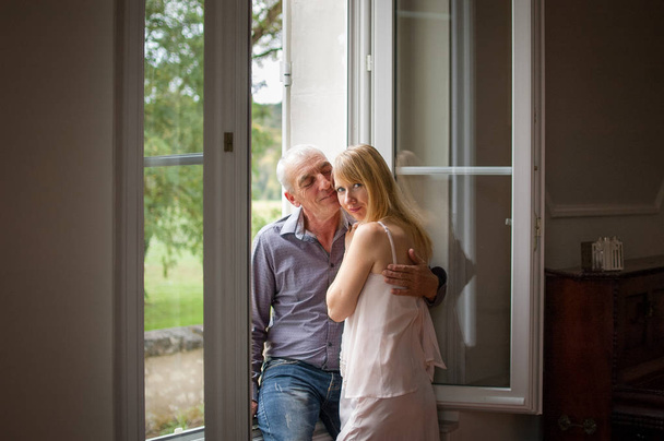 Senior man omhelst zijn jonge Blond vrouw in de buurt van het raam. Een vrouw die naar de camera kijkt. Psychologie van relaties Concept. - Foto, afbeelding