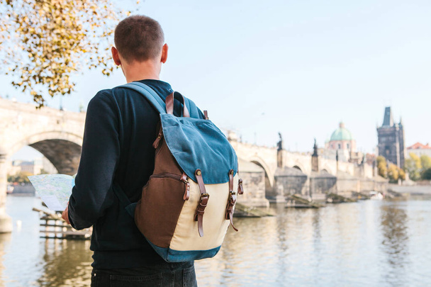 Een jonge toeristische man met een rugzak die naast de rivier Vltava in Praag kijkt naar de kaart en de architectuur van de stad bewondert. Charles Bridge is in de buurt. Rondreis door Tsjechië. - Foto, afbeelding