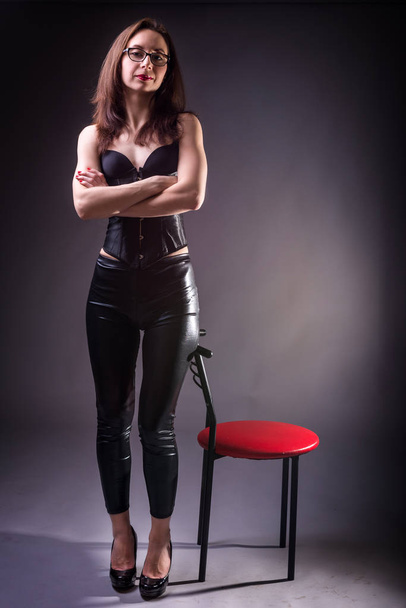Sexy young woman in corset and latex pants posing near a chair on a gray background. - 写真・画像
