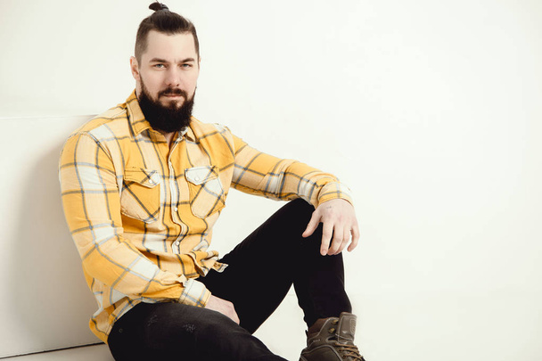 Fashion portrait of young bearded man in yellow plaid shirt and black jeans sitting over white background. - Фото, зображення