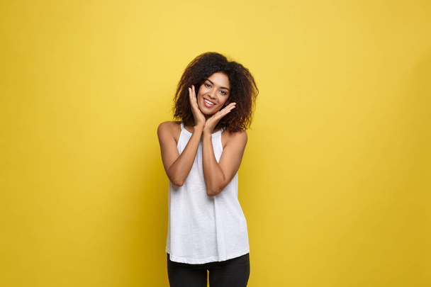 Sonriente hermosa joven afroamericana en camiseta blanca posando con las manos en la barbilla. Estudio filmado sobre fondo amarillo. Copiar espacio
. - Foto, imagen