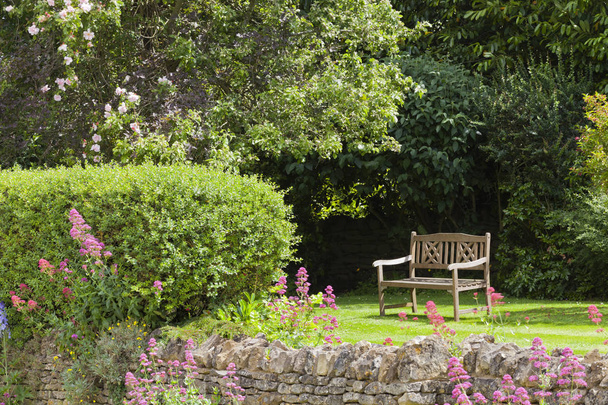 Afgelegen houten bankje in een Engelse zomertuin met roze rosebush, cottage bloemen, struiken, door rotsachtige stenen muur . - Foto, afbeelding