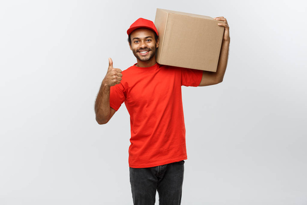 Koncepcja dostawy - portret szczęśliwy African American dostawy człowiek posiadający pakiet pole i pokazuje thumps. Na białym tle na studio szary tło. Miejsce. - Zdjęcie, obraz