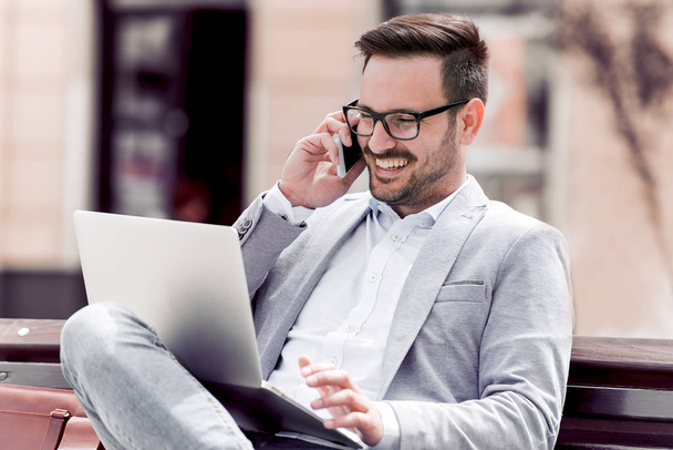 schöner junger Geschäftsmann sitzt auf einer Bank mit seinem Laptop auf einer Straße neben einem Park, während er mit seinem Handy telefoniert - Foto, Bild