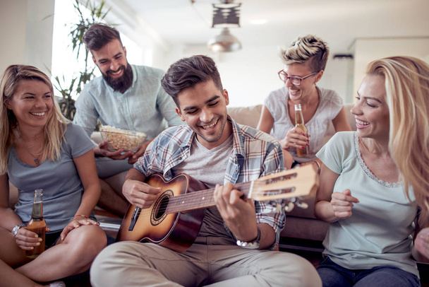 Groep vrienden met plezier in de woonkamer. Gelukkige jonge mensen gitaar spelen, zingen en lachen samen. - Foto, afbeelding