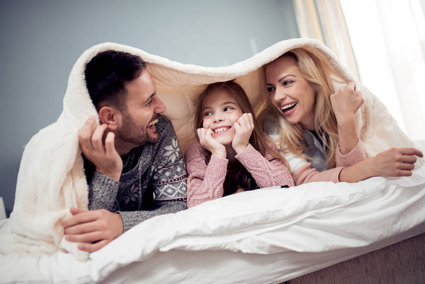 Mamma, papà e figlia sdraiati sotto la coperta, guardando film e divertendosi
. - Foto, immagini