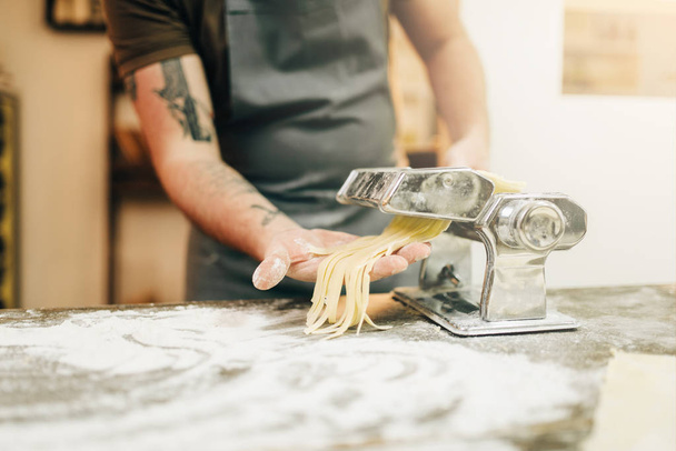 σεφ στην ποδιά εργασίας με ζύμη από μηχανή ζυμαρικών στο τραπέζι της κουζίνας ξύλινα - Φωτογραφία, εικόνα