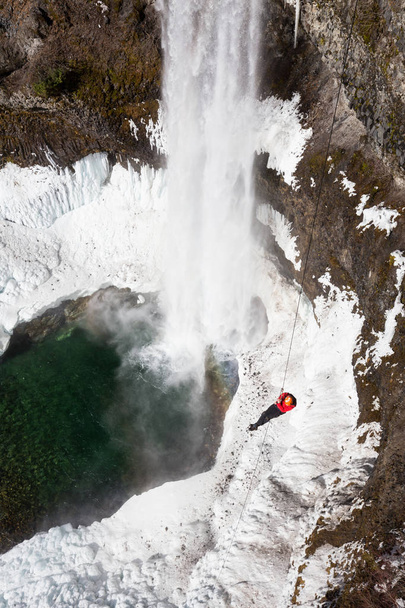 Adventurous young man is rappeling down a steep cliff beside a big and beautiful waterfall. Taken in Brandywine Falls, near Whister and Squamish, North of Vancouver, BC, Canada. - Foto, afbeelding