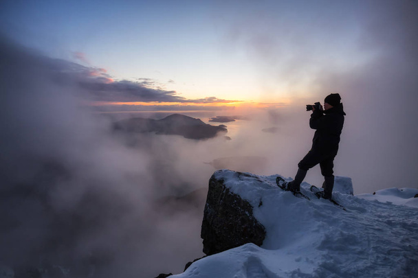 Adventurous man on the side of a steep cliff is taking pictures of the beautiful view during a vibrant sunset. Taken on top of St Mark's Peak, North of Vancouver, BC, Canada. - Foto, Imagen