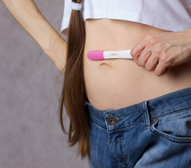 Jovem entre 30 e 40 anos de idade vestida com jeans mantém o teste de gravidez perto da barriga. Fechar.
 - Foto, Imagem