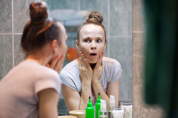 Девушка в возрасте от 30 до 40 лет наблюдает за своей кожей лица перед зеркалом в ванной комнате
. - Фото, изображение