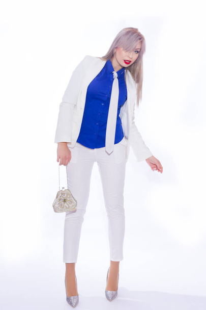 Studio portrait in full length of a beautiful young blond woman with long hair in a white pants suit. A girl in silvery shoes with high heels, in her hands she holds a small silver handbag - Photo, image