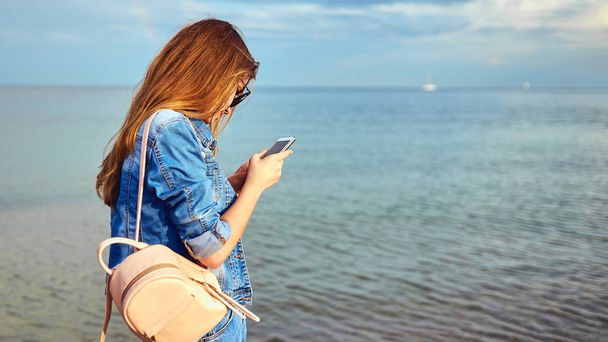A tourist girl walks along the seashore with a backpack, in junks and sunglasses. With a mobile phone, takes photos on a smartphone. Selfie - Foto, afbeelding