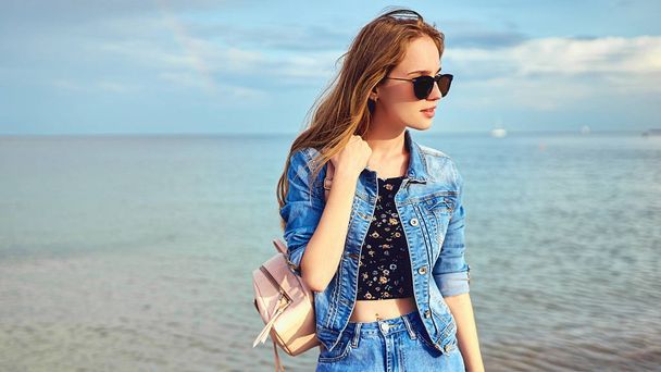 A tourist girl walks along the seashore with a backpack, in junks and sunglasses. With a mobile phone, takes photos on a smartphone. Selfie - Foto, Bild