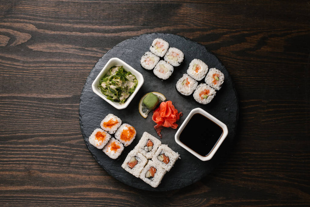 Vista superior de diferentes tipos de sushi maki servido em placa de pedra escura com gengibre marinado, wasabi, algas marinhas e molho de soja
 - Foto, Imagem
