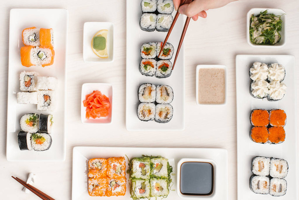 Widok z góry męskich rąk pałeczkami, wybór różnych rodzajów maki sushi serwowane białe płytki na beżowy powierzchni drewnianych - Zdjęcie, obraz