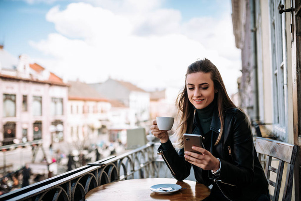 Młoda kobieta, picia kawy i przy użyciu telefonu siedząc na balkonie z widokiem na miasto.  - Zdjęcie, obraz