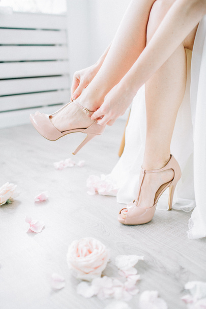 In einem hellen Raum mit rosafarbenen Blütenknospen zieht sich die junge Frau rosafarbene High Heels an. Brautmodenkonzept. - Foto, Bild